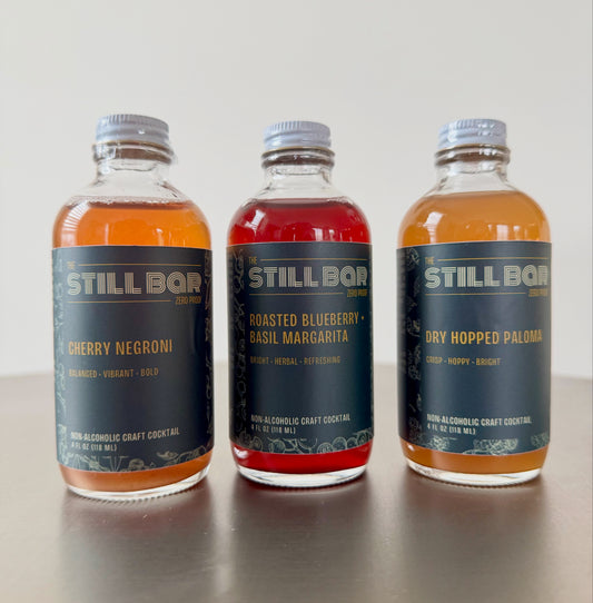 Three bottles of The Still Bar non-alcoholic craft cocktails on a reflective surface with a light gray background.