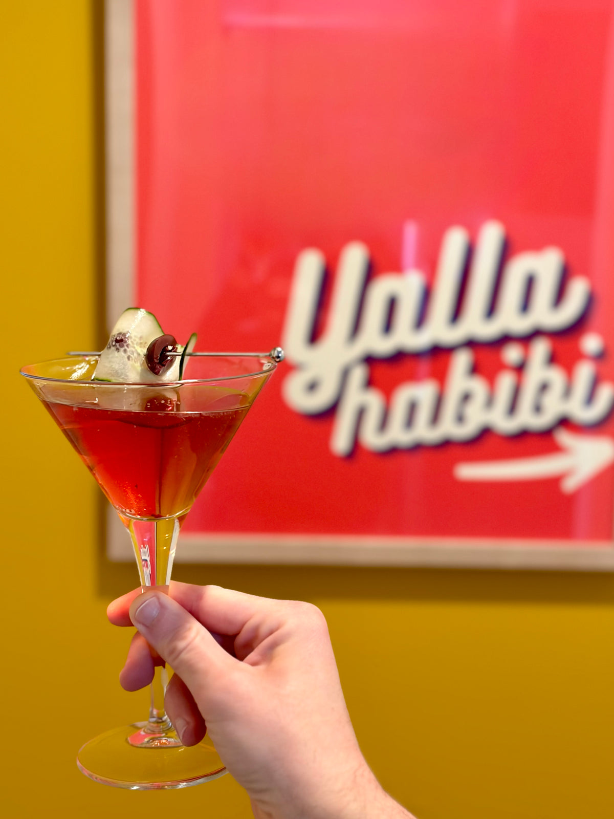 Hand holding a Yalla Martini in front of wall art that says "Yalla habibi"