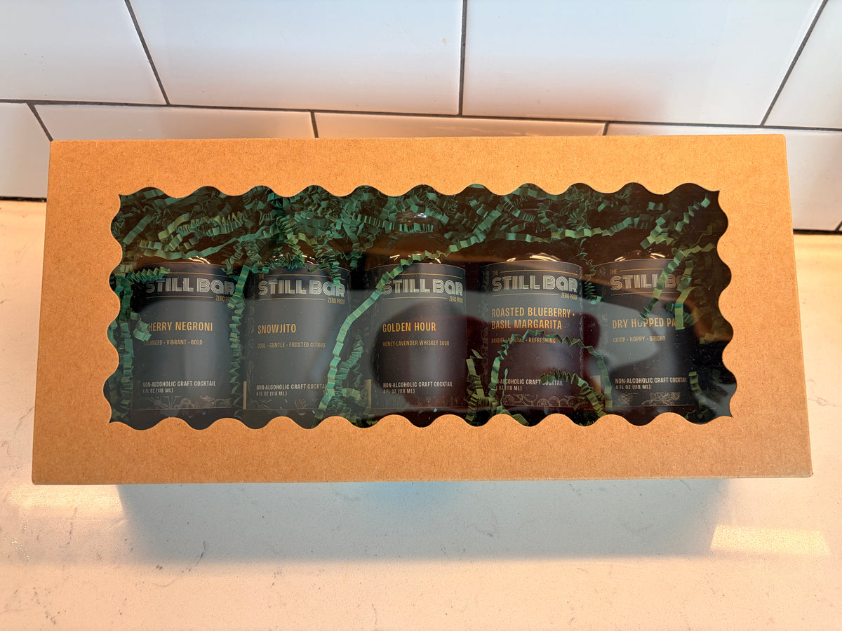 A gift box containing five dark glass bottles from the brand The Still Bar, visible through a decorative green leaf-patterned window cutout on a tan cardboard box.