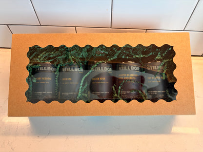 A gift box containing five dark glass bottles from the brand The Still Bar, visible through a decorative green leaf-patterned window cutout on a tan cardboard box.