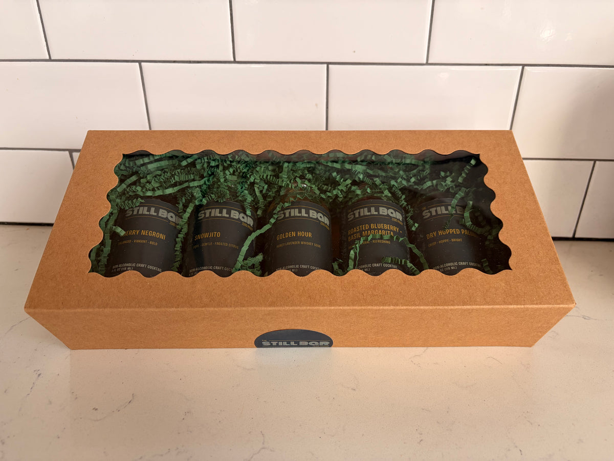 A kraft paper gift box with a clear window displaying five dark bottles of The Still Bar's cocktails on green paper filler, set on a light countertop against a white subway tile backsplash.