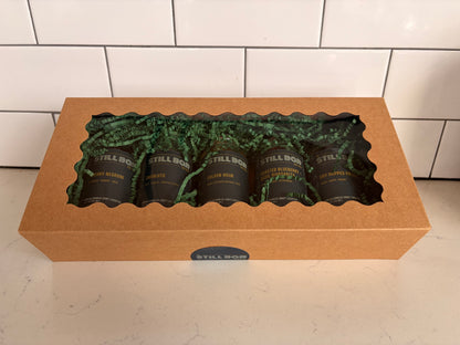 A kraft paper gift box with a clear window displaying five dark bottles of The Still Bar's cocktails on green paper filler, set on a light countertop against a white subway tile backsplash.