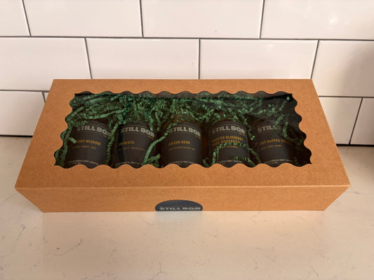 A kraft paper gift box with a clear window displaying five dark bottles of The Still Bar's cocktails on green paper filler, set on a light countertop against a white subway tile backsplash.