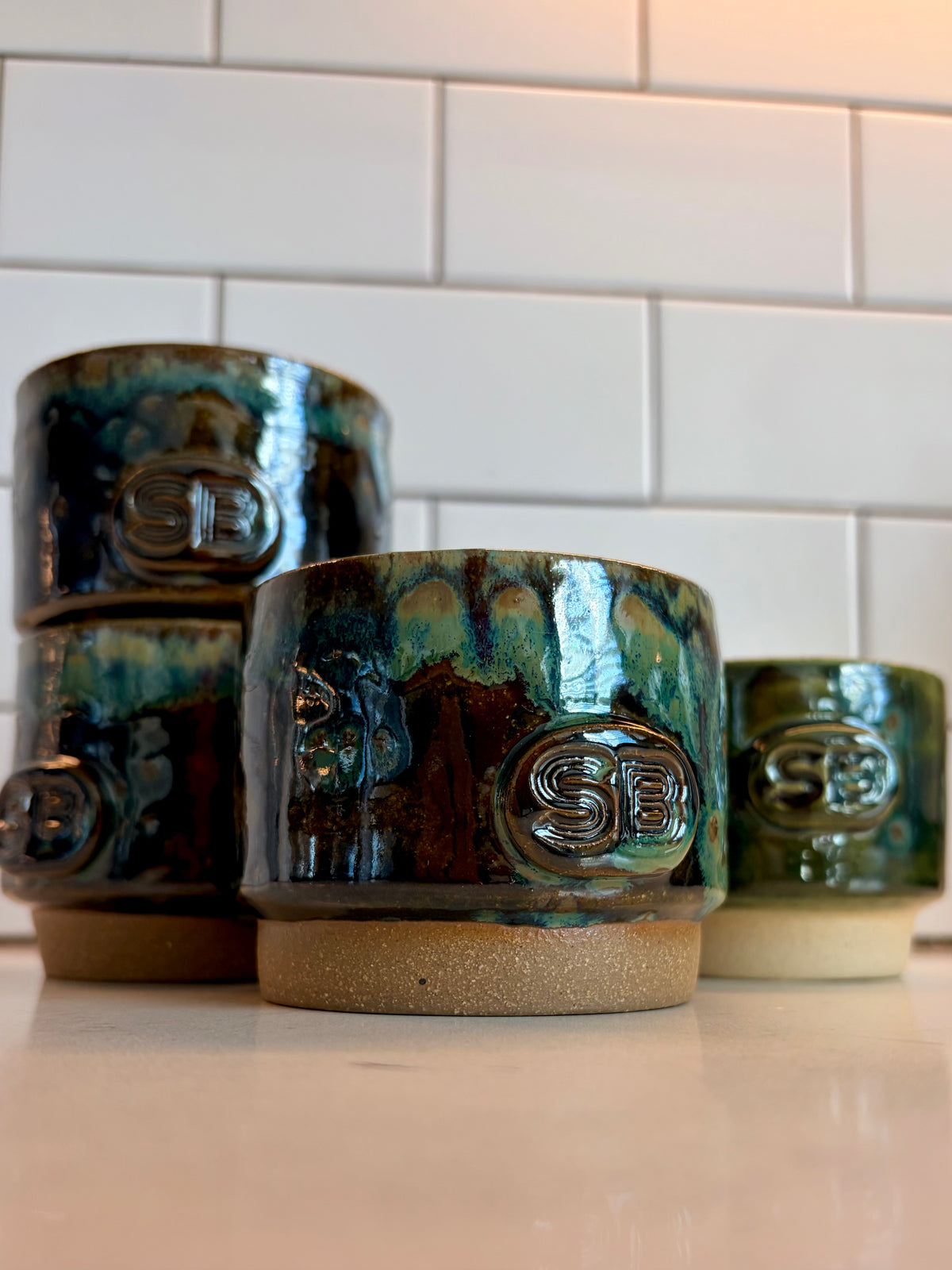 A set of handmade ceramic cups with a glossy teal, green, and brown glaze, featuring an embossed 'SB' monogram on the front and a speckled tan base, arranged on a countertop against a white subway tile backsplash.