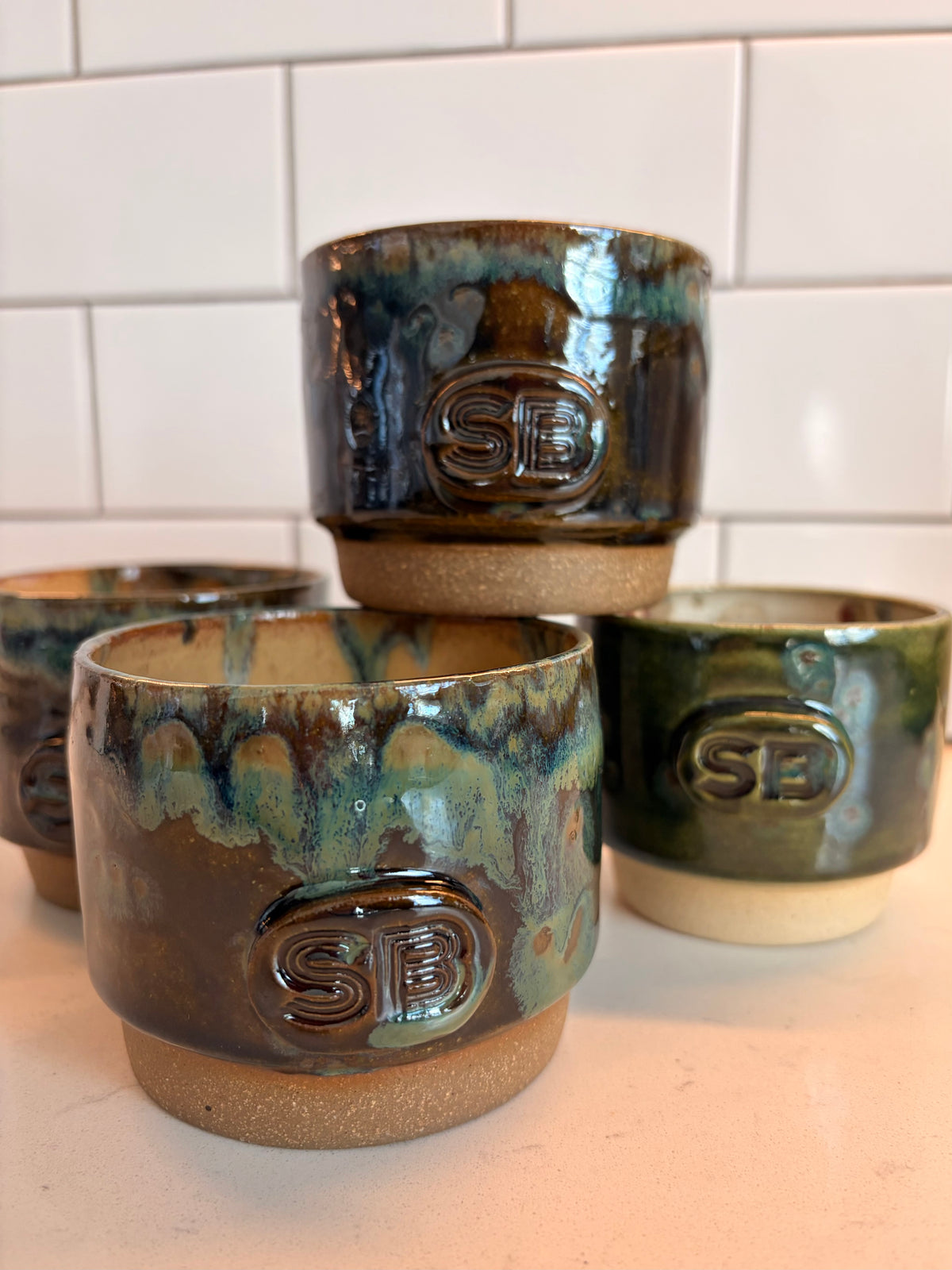 Four handmade ceramic tumblers with a glossy brown and green glaze, stacked and arranged on a countertop against a white subway tile backsplash. Each cup features an embossed monogram logo reading 'SB'.