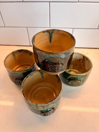Four handmade ceramic cups with a rustic green and brown glaze, arranged on a white countertop against a white subway tile backsplash.