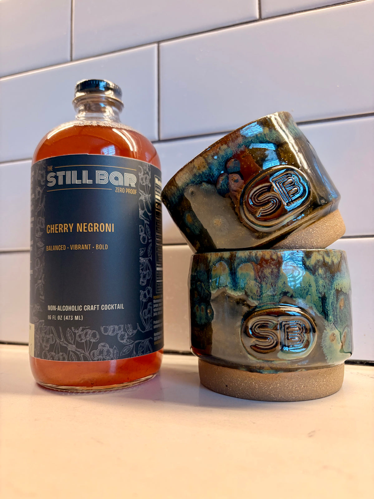 A bottle of Still Bar Zero Proof Cherry Negroni non-alcoholic craft cocktail stands next to two stacked ceramic tumblers with a blue-green glazed finish and an embossed SB logo, set against a white subway tile background.