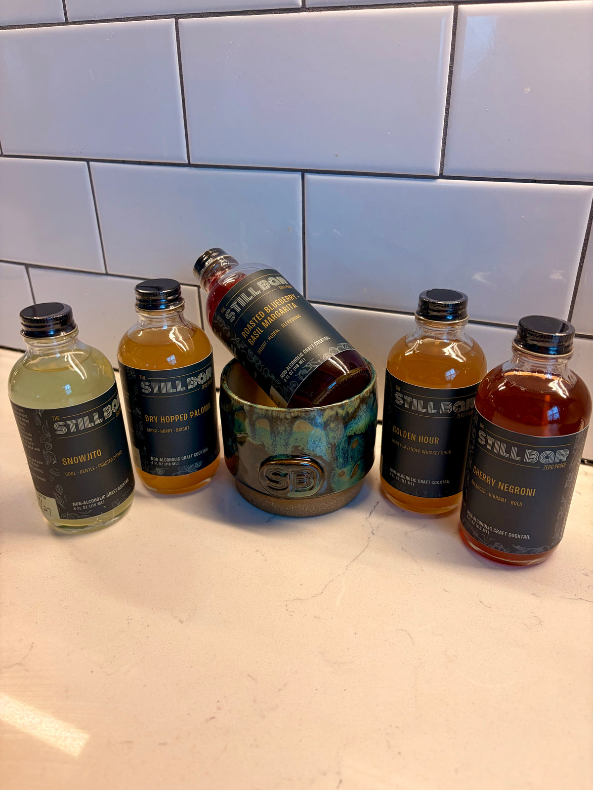 Six individual bottles of The Still Bar craft cocktails arranged on a white countertop in front of a white subway tile wall. A central rustic ceramic cup holds one bottle, while the other bottles stand upright around it.