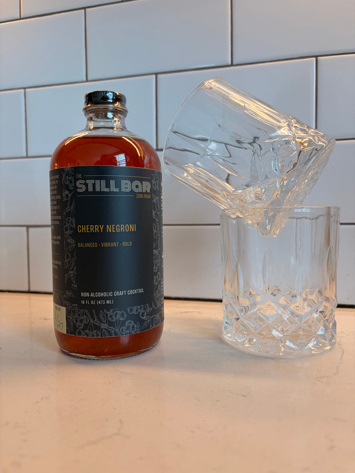 A bottle of The Still Bar Cherry Negroni non-alcoholic craft cocktail stands on a countertop next to two clear cut-glass tumblers, with a white subway tile backsplash behind it.