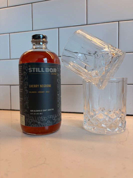 A bottle of The Still Bar Cherry Negroni non-alcoholic craft cocktail stands on a countertop next to two clear cut-glass tumblers, with a white subway tile backsplash behind it.