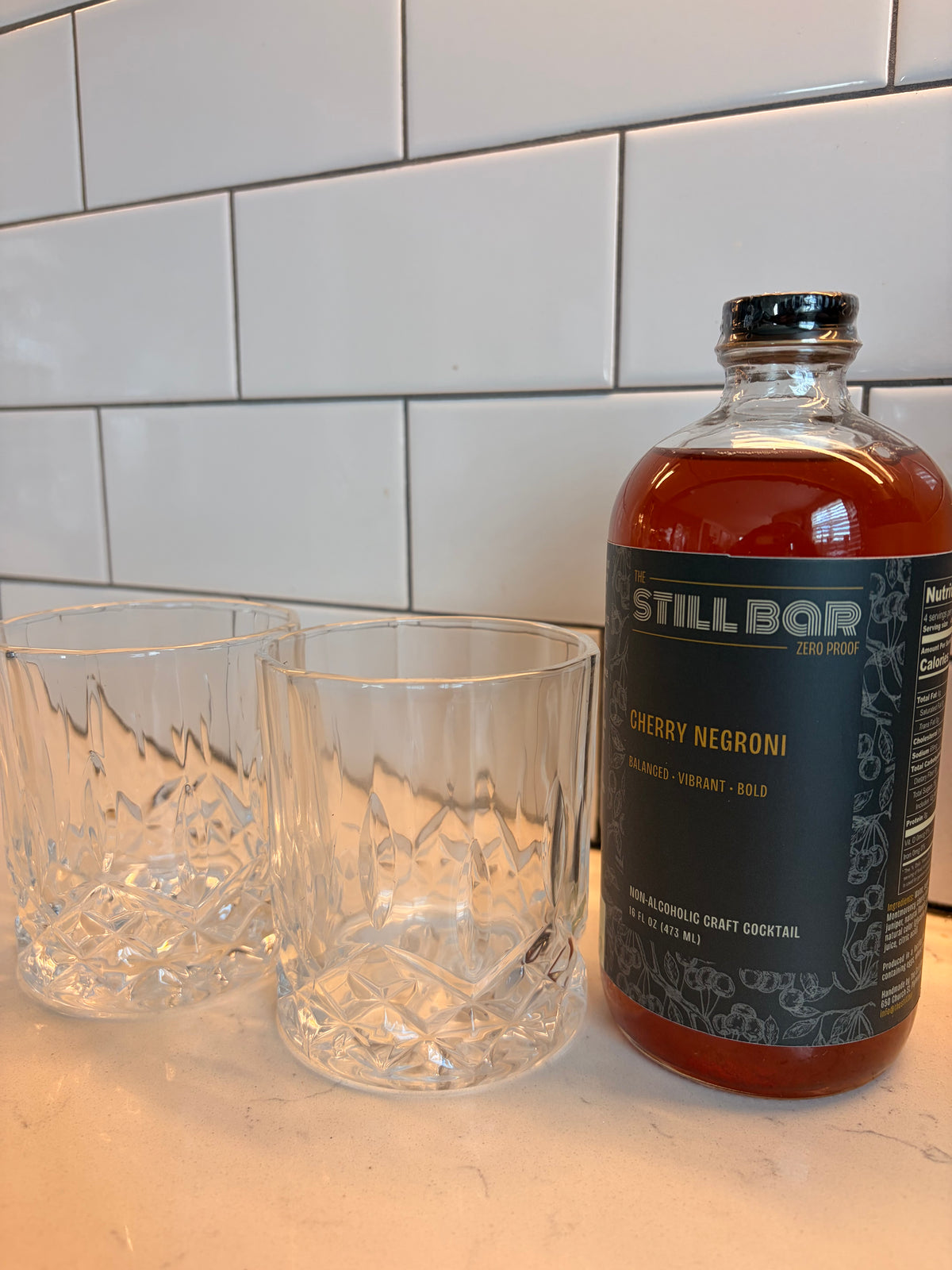 A bottle of Still Bar Cherry Negroni alcoholic craft cocktail next to two clear cut-glass rocks tumblers, set on a light countertop against a white subway tile backsplash.