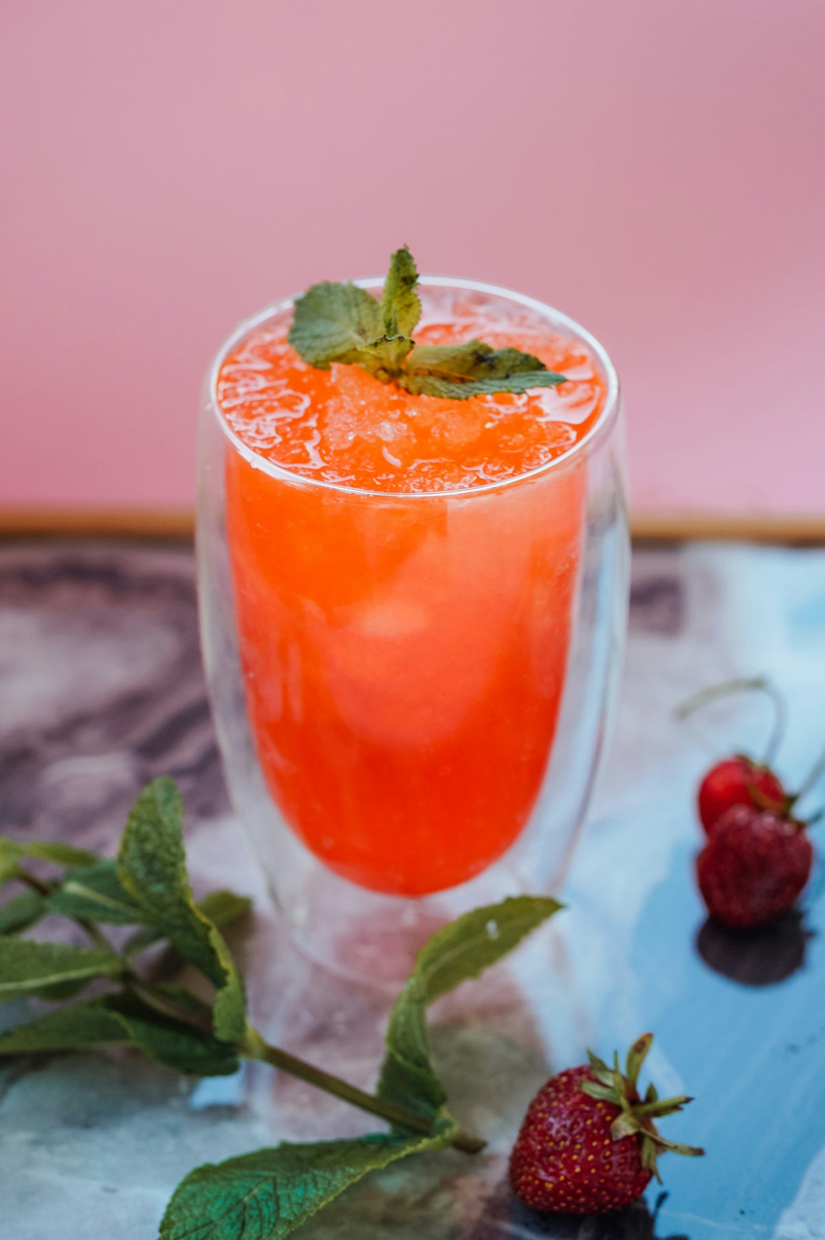Orange cocktail in a clear glass with strawberries and mint leaves on a pink background